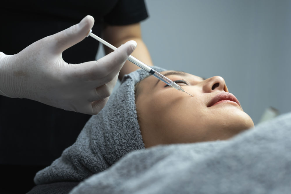 Close up beautician wearing hygiene gloves injecting filler on woman cheek for facial treatment to female Asian client in beauty clinic